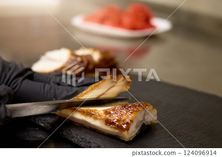 A chef with black gloves skillfully slices tender roasted meat on a slate board. Fresh ingredients can be seen in the background, enhancing the culinary atmosphere 124096914