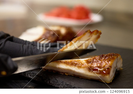 A skilled chef slices through crispy pork in a modern kitchen. The juicy meat is complemented by vibrant garnishes placed on a white plate, showcasing culinary precision and artistry A skilled chef slices through crispy pork in a modern kitchen. The juicy meat is complemented by vibrant garnishes placed on a white plate, showcasing culinary precision and artistry 124096915