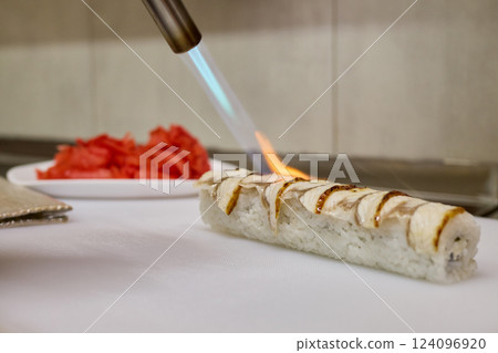 A chef uses a torch to skillfully flame-sear sushi in a restaurant kitchen. Fresh slices of fish are displayed on a plate nearby, emphasizing the focus on quality ingredients 124096920