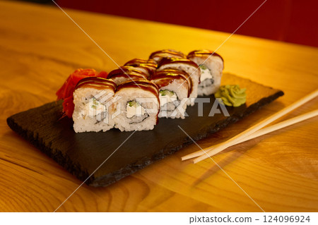 Fresh sushi rolls filled with cream and vegetables are stylishly presented on a black stone plate. Vibrant wasabi and pickled ginger accompany the dish in a cozy dining setting 124096924