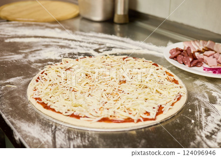 A pizza base topped with rich tomato sauce and generously sprinkled with shredded cheese sits on a stainless steel countertop in a lively kitchen 124096946