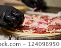 Hands wearing black gloves delicately arrange slices of meat and onion on a pizza base in a bustling kitchen, creating a flavorful dish for eager customers 124096950