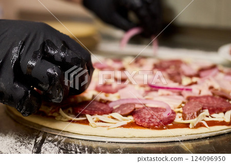 Hands wearing black gloves delicately arrange slices of meat and onion on a pizza base in a bustling kitchen, creating a flavorful dish for eager customers Hands wearing black gloves delicately arrange slices of meat and onion on a pizza base in a bustling kitchen, creating a flavorful dish for eager customers 124096950