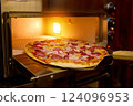 A freshly baked pizza with pepperoni, ham, and onions is being carefully pulled from the oven in a warm, inviting kitchen environment during the evening 124096953