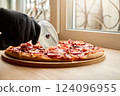 A hand wearing a black glove uses a pizza cutter to slice a pepperoni pizza on a wooden board. The warm afternoon light streams through a nearby window, creating a homey atmosphere 124096955