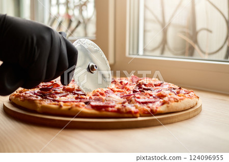 A hand wearing a black glove uses a pizza cutter to slice a pepperoni pizza on a wooden board. The warm afternoon light streams through a nearby window, creating a homey atmosphere 124096955