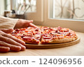 A person is gently placing a deliciously topped pizza on a wooden board by a sunny window. The warm colors create a welcoming atmosphere for enjoying a meal 124096956
