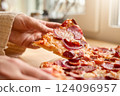 A hand reaches for a slice of pepperoni pizza on a wooden table in a bright kitchen. The pizza is loaded with toppings, showcasing a mouthwatering feast perfect for lunch or dinner 124096957