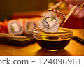 A hand holds a chopstick lifting a sushi roll filled with cream cheese and vegetables, ready to dip into soy sauce at a warm and inviting restaurant setting 124096961