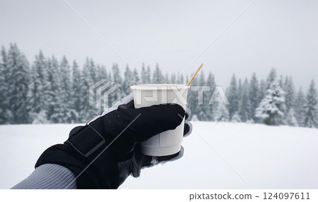 Person hand holding a cup of hot mulled wine or coffee outdoors on the top of the mountain in the winter day with snow falling. Wonderful white covered fir forest on the background. Person hand holding a cup of hot mulled wine or coffee outdoors on the top of the mountain in the winter day with snow falling. Wonderful white covered fir forest on the background. 124097611