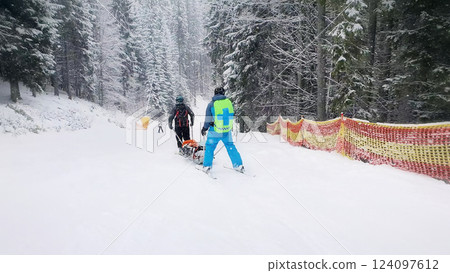 Mountain rescuers transporting an injured skier, going downhill after first aid. Slope rescue, ski patrol service evacuating the victim on a special sledge. Bukovel ski resort, ukraine carpathians. 124097612