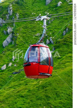 Scenic View of a Modern Red Cable Car Cabin Against a Stunning Green Mountainside Landscape Scenic View of a Modern Red Cable Car Cabin Against a Stunning Green Mountainside Landscape 124098984
