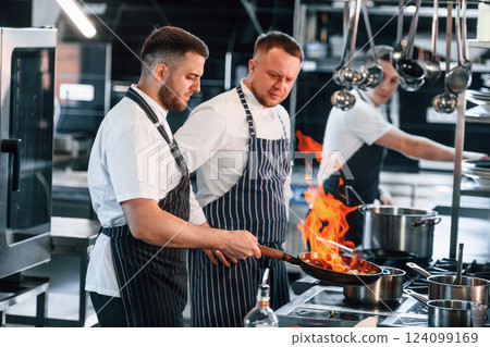 Flaming the meal on frying pan. Kitchen workers is together preparing the food Flaming the meal on frying pan. Kitchen workers is together preparing the food 124099169