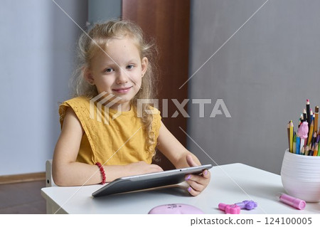 A happy little girl is playing with a tablet. Uses the gadget for learning and communication. Enjoys interesting video or photo content on social networks. 124100005