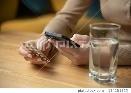 Man checks medication instructions while holding a phone and glass of water at a cozy table in the afternoon 124101225