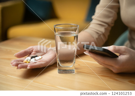 Individual examines medication instructions on smartphone while holding pills and water at home during afternoon 124101253