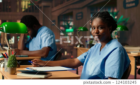 African american med school pupil studies pharmacology notes in a library, preparing for exams and advancing their healthcare career. Girl in scrubs works hard on medical school success. Camera B. African american med school pupil studies pharmacology notes in a library, preparing for exams and advancing their healthcare career. Girl in scrubs works hard on medical school success. Camera B. 124103245