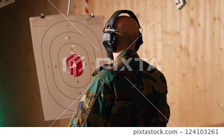 Happy military personnel in firing range shoots targets with handgun, satisfied after achieving positive result. Joyous soldier training with firearm exults after accurately hitting bullseye, camera B 124103261