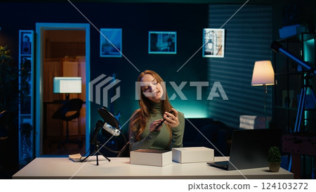 Gen Z content creator filming technology review of newly released smartphone, unpacking it and presenting specifications to audience. Internet star showing cellphone to fans from studio, camera B 124103272