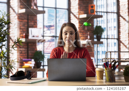 Mature woman smiling and waving during a video call at her home office desk, showcasing modern technology and online communication in a digital and wireless lifestyle. Web conference. 124103287