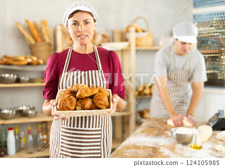 Portrait of happy female baker working in bakehouse, holding tray with fresh bakery goods on kitchen Portrait of happy female baker working in bakehouse, holding tray with fresh bakery goods on kitchen 124105985