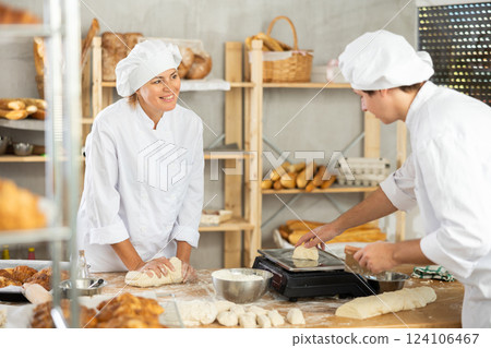 Two bakers in white uniform. Skilled female baker working with young male assistant in bakery, kneading raw dough, cutting and weighing portioned pieces for baking Two bakers in white uniform. Skilled female baker working with young male assistant in bakery, kneading raw dough, cutting and weighing portioned pieces for baking 124106467