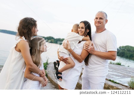 Family of five is standing on pier by the water, with woman holding baby. Atmosphere is warm and friendly Family of five is standing on pier by the water, with woman holding baby. Atmosphere is warm and friendly 124112211