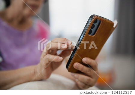 Elderly person using smartphone with a brown leather case while sitting indoors, close-up of hand and mobile device in a cozy environment 124112537