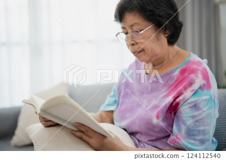 Senior Asian Woman Enjoying a Peaceful Reading Session on a Cozy Sofa in a Bright Living Room with Natural Light and Minimalist Decor 124112540
