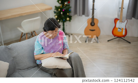 Elderly Asian woman reading a book on a cozy sofa in a decorated living room with a small Christmas tree and musical instruments in the background Elderly Asian woman reading a book on a cozy sofa in a decorated living room with a small Christmas tree and musical instruments in the background 124112542