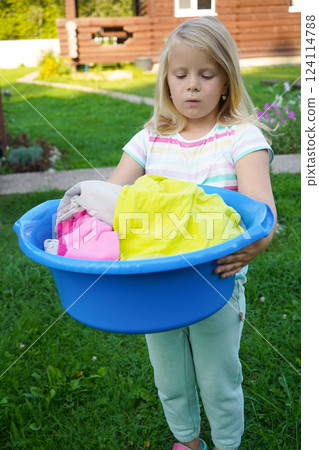 A six-year-old girl, holding a plastic tub, stands near the clothesline, enjoying the fresh outdoor air. A six-year-old girl, holding a plastic tub, stands near the clothesline, enjoying the fresh outdoor air. 124114788