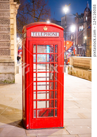 A traditional red telephone box stands illuminated against the night sky in London, showcasing the city's vibrant ambiance. Nearby streets are aglow with city lights and a bustling atmosphere. A traditional red telephone box stands illuminated against the night sky in London, showcasing the city's vibrant ambiance. Nearby streets are aglow with city lights and a bustling atmosphere. 124116088