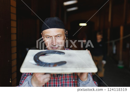 European busy blacksmith inspects horseshoe made for horse in stable 124118319
