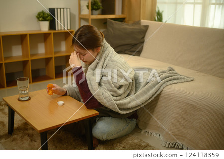 Sick young woman wrapped in a warm blanket and taking medicine for recovery 124118359