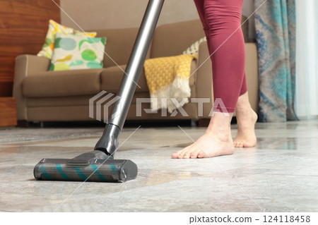 Modern upright cordless vacuum cleaner with dust light. Woman cleaning living room near sofa.  124118458