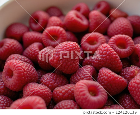 A close-up image of fresh, ripe raspberries in a white bowl. The berries are vibrant red, showcasing their natural texture and juicy appearance.  124120139