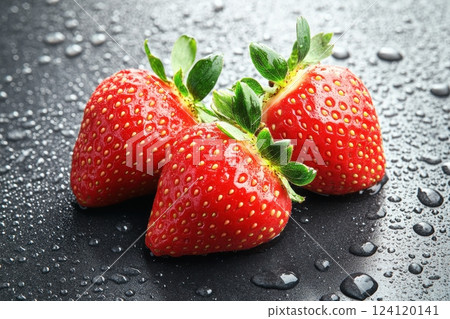 A close-up image of three fresh, ripe strawberries on a dark, wet surface. The strawberries are vibrant red with visible seeds and green leaves still attached.  124120141