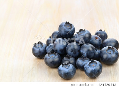 A close-up image of fresh, ripe blueberries scattered on a light wooden surface. The berries are a deep blue-purple color, showcasing their natural bloom and plump, round shape.  124120148