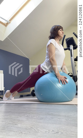 Woman in 50's doing pilates on fitness ball at home. Sports equipment and room for sports. Health and exercise. Woman in 50's doing pilates on fitness ball at home. Sports equipment and room for sports. Health and exercise. 124120861