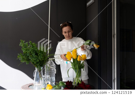Woman arranging vibrant yellow tulips in a modern indoor setting with greenery on display 124121519
