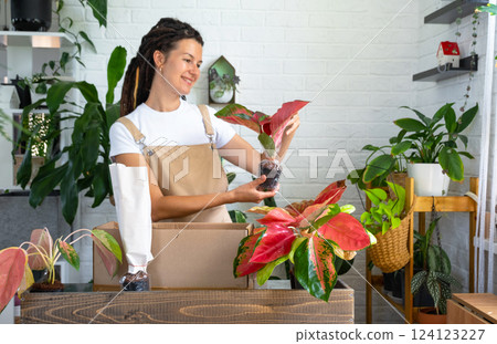 Woman unpacks a box with a parcel of potted flowers of red aglaonem - order wrapped tropical plants, delivery. Sale, safe shipment of plants from the store, packaging. Flower shop, home business 124123227