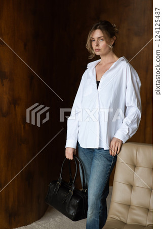 A girl in a white shirt and blue jeans is posing with a black bag. 124125487