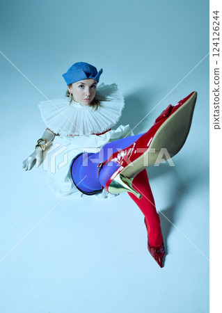View from above of young woman, dressed red-blue tights, corset with collar lifting leg to camera against studio background. 124126244