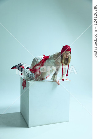 Young woman in white lace dress with red accents crawling on white cube, gazing intensely. Concept of surreal fashion Young woman in white lace dress with red accents crawling on white cube, gazing intensely. Concept of surreal fashion 124126296