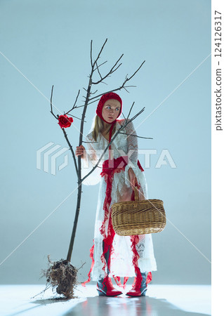 Young woman in white lace dress, red accents, and knitted hat holding basket and standing beside uprooted tree with single red rose 124126317