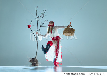 Woman in fluid motion, holding basket, posed beside bare tree with blooming rose, symbolizes fragile resilience and change. Woman in fluid motion, holding basket, posed beside bare tree with blooming rose, symbolizes fragile resilience and change. 124126318