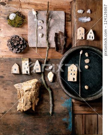 Flat lay creative design rustic composition with miniature ceramic houses, pebbles, shell, moss, pine cone, feather, twigs on wooden background. Natural materials arrangement. 124126867