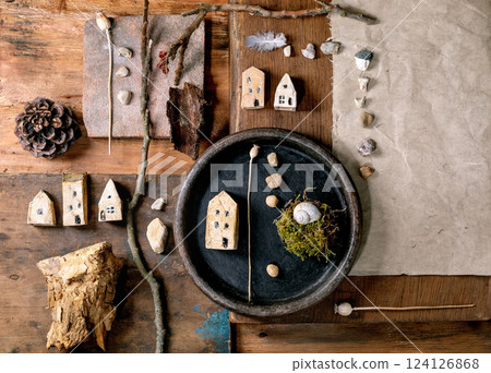 Flat lay creative design rustic composition with miniature ceramic houses, pebbles, shell, moss, pine cone, feather, twigs on wooden background. Natural materials arrangement. 124126868