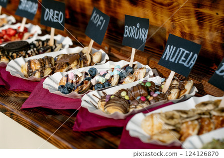 A variety of sweet and savory pancakes topped with Oreos, Nutella, bananas, and more. Each serving is labeled and presented in a food market setting, offering diverse flavors. 124126870