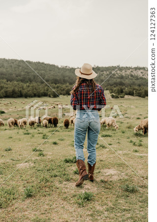 A woman wearing a hat and boots walks through a field of sheep 124127363